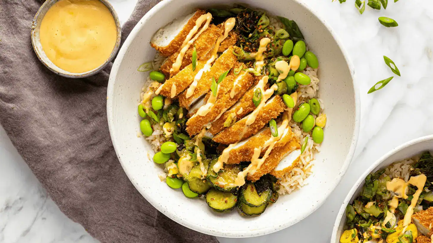 Overhead shot of two appetizing Crispy Chicken Katsu Bowls with brown rice, edamame, and a creamy sauce, presented on a white marble surface.